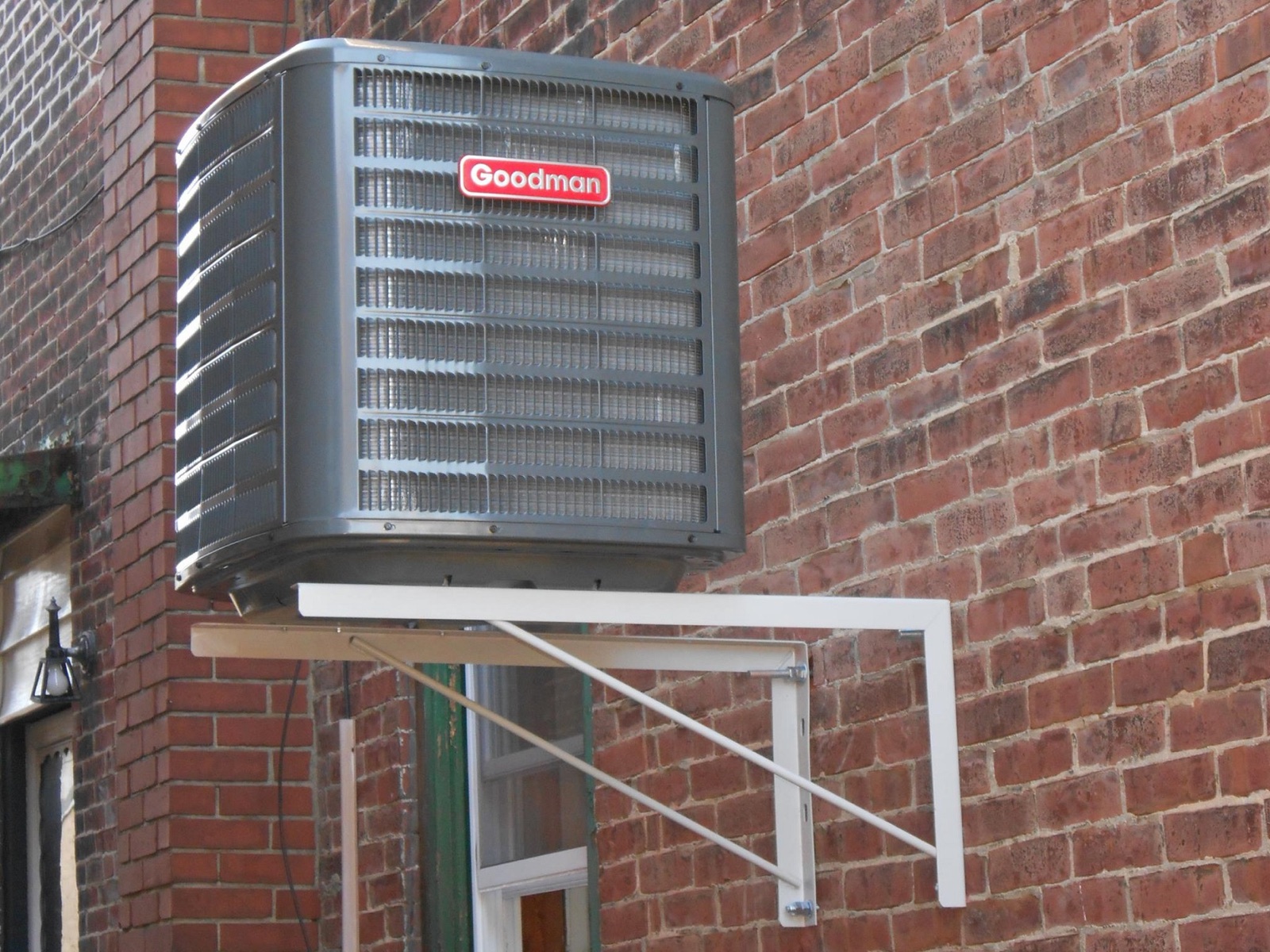 Goodman AC condenser mounted on a wall bracket on a brick building
